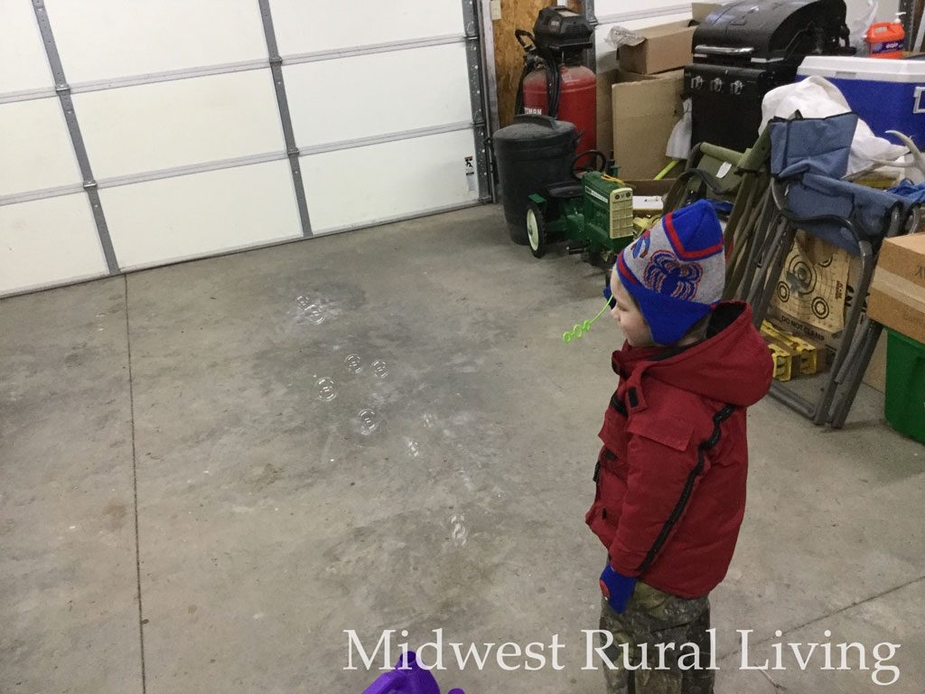 Colt is blowing bubbles in the garage.  It is cold and rainy outside so he is has is insulated coat, bibs and Spiderman hat on.  